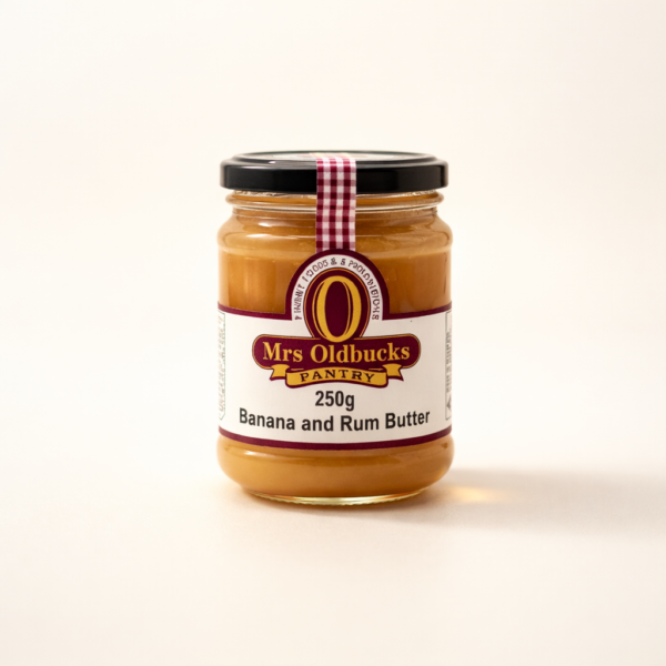 Mrs Oldbucks Pantry Banana Rum Butter 250g jar of Mrs Oldbucks Pantry Banana Rum Butter on a warm ivory background, showing smooth golden banana butter with subtle richness in a clear glass jar with black lid and burgundy label.