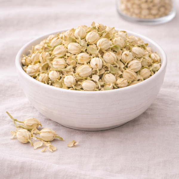 Mrs Olbucks Pantry Jasmine Blossom White ceramic bowl filled with dried jasmine buds and petals, styled on soft neutral linen. A few loose buds rest in front of the bowl, with a small glass jar of jasmine buds softly blurred in the background. Soft, natural light highlights the pale cream and green tones of the blossoms.