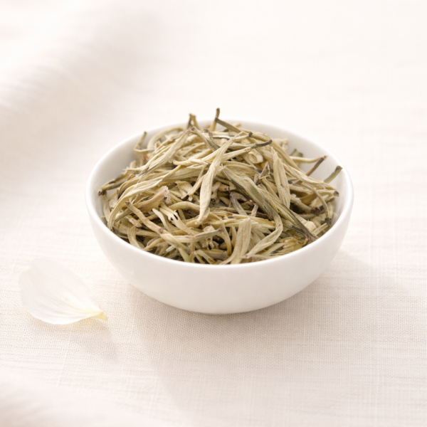 Mrs Oldbucks Pantry Silver Needles loose leaf white tea in a small white ceramic bowl on soft ivory linen. The long, pale silver buds with fine downy tips are softly illuminated from the top left, with a single delicate white petal resting beside the bowl in a light, minimalist studio setting.