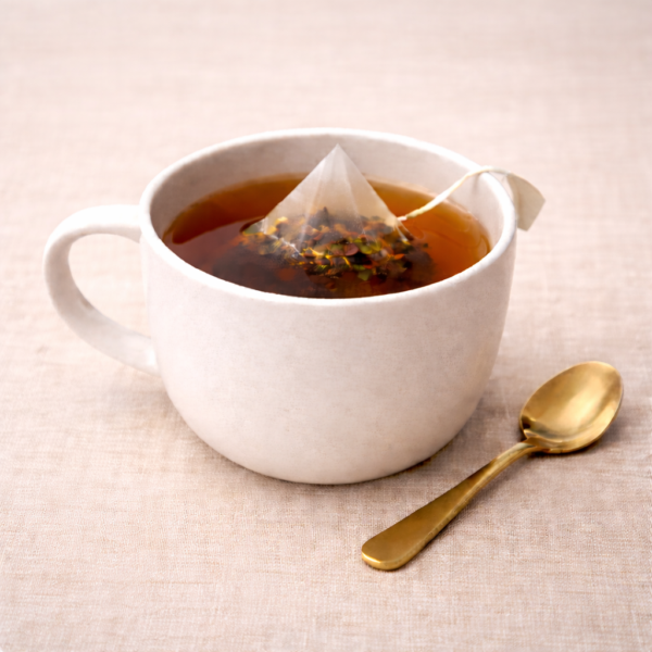Mrs Oldbucks Pantry Punjabi Chai Pyramid Teabags steeping in a white ceramic cup with pale golden herbal infusion, set on warm neutral linen with a brass teaspoon beside the cup in soft top-left lighting.
