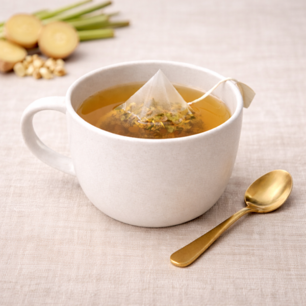 Mrs Oldbukcs Pantry Lemongrass and Ginger pyramid tea bag steeping in a white ceramic cup with light golden herbal infusion, set on warm neutral linen with a brass teaspoon beside the cup in soft top-left lighting.