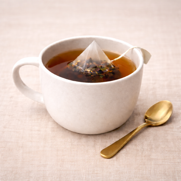 Mrs Oldbucks Pantry Melbourne Breakfast Pyramid Teabags steeping in a white ceramic cup with warm amber liquor, set on neutral linen with a brass teaspoon beside the cup in soft top-left lighting.