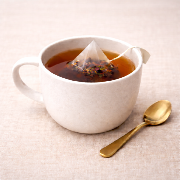 Mrs Oldbucks Pantry Springtime Grey Pyramid Teabags steeping in a white ceramic cup with pale golden herbal infusion, set on warm neutral linen with a brass teaspoon beside the cup in soft top-left lighting.