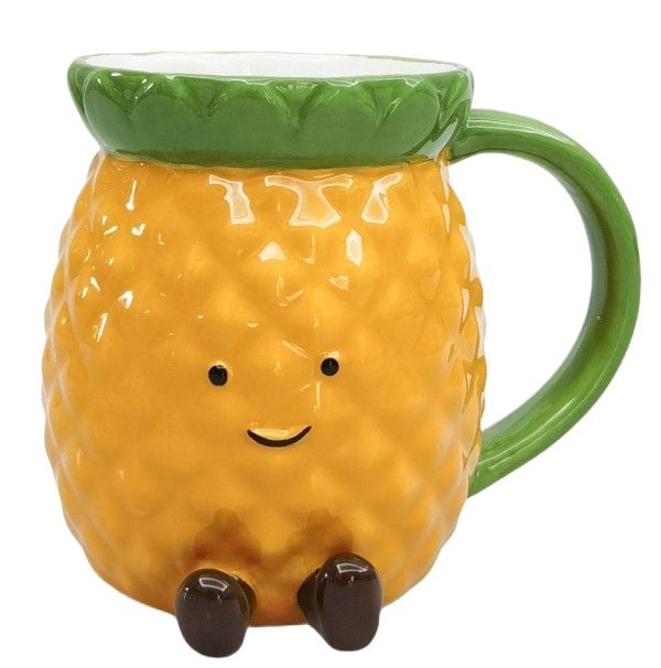 Mrs Oldbucks Pantry Penny Pineapple Mug Pineapple-shaped ceramic mug with textured golden body, green leaf rim and handle, smiling face and small brown feet on a white background.